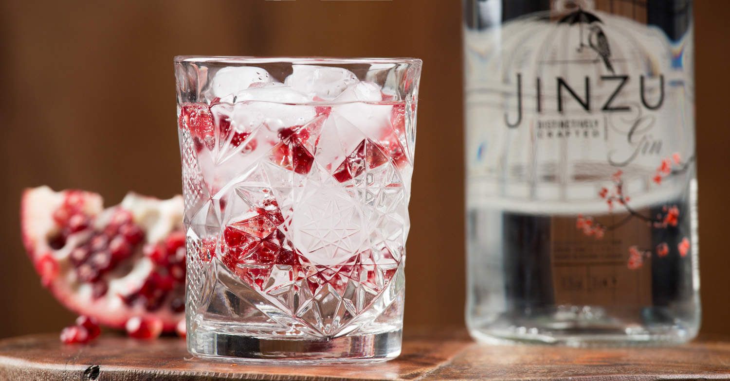 Tumbler Glas mit einem Gin Tonic, Eis und roten Beeren. Im Hintergrund eine Flasche Jinzu Gin.