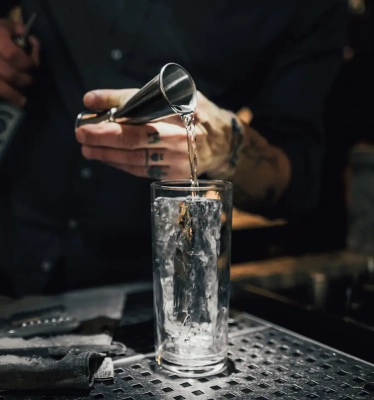 Barkeeper der Gin in einem Jigger in ein mit Eis gefülltes Longdrinkglas gießt.
