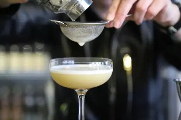 Gelber Gin Cocktail mit Schaumkrone, der durch ein Sieb abgeseiht wird. 